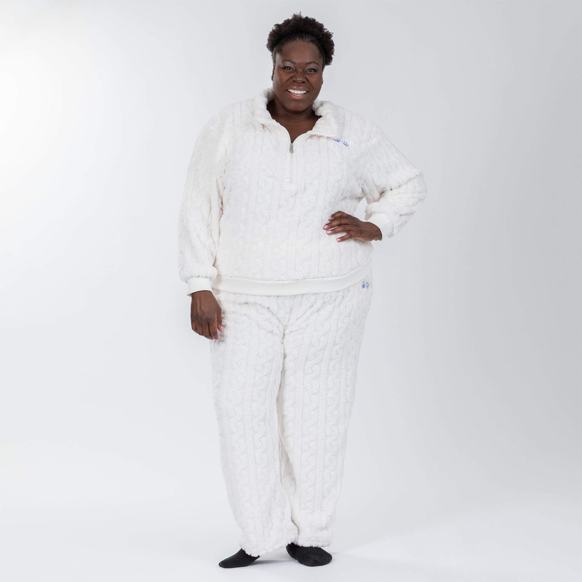 Woman wearing a white, fleece, cable-knit, quarter-zip, long-sleeve shirt with two light blue paw prints embroidered beneath the lapel. Her pants are part of the matching set and are also white with a cable-knit pattern and two light blue paw prints embroidered on the lefthand pocket.  Extended and plus size available. 