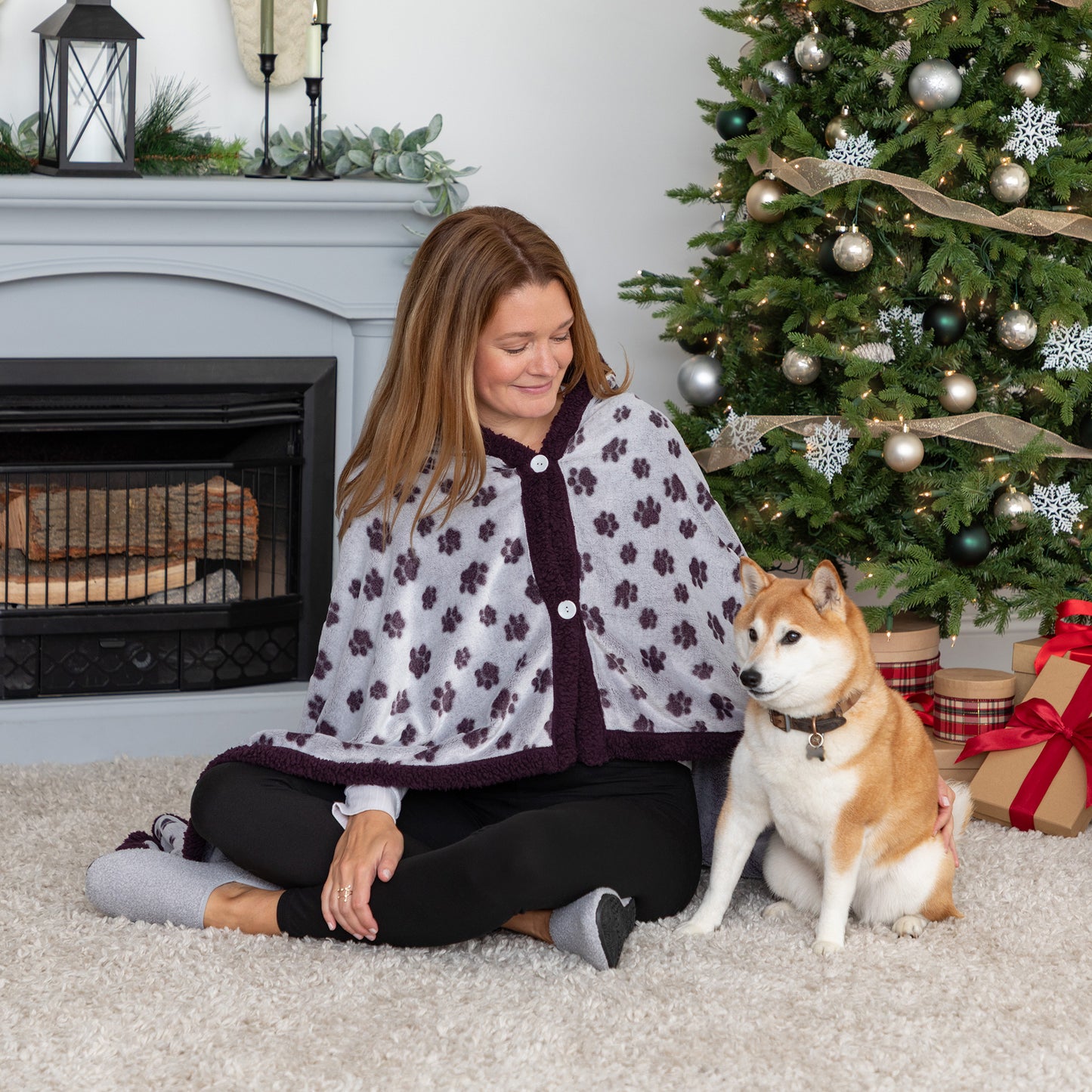 Woman in a floral cape sitting on the floor with a dog in front of a Christmas tree.