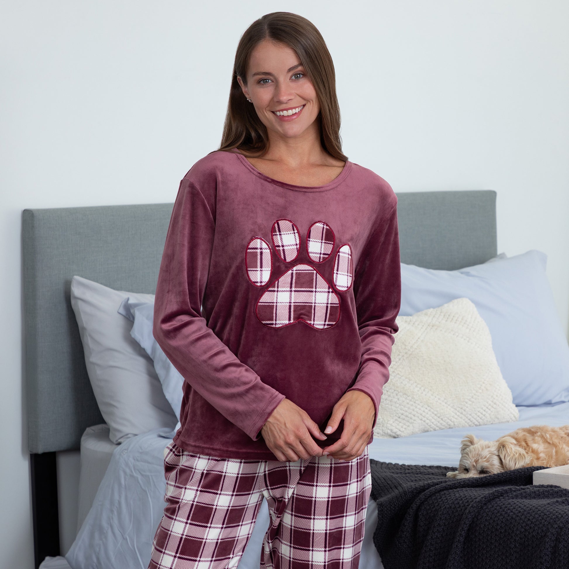 Woman standing in cozy bedroom setting while her pet dog snuggles up in the bed behind her. She is wearing a velour pajama set— the top is a maroon long-sleeve shirt with a large plaid paw print. The pants are the same white and maroon plaid pattern as the paw print on the shirt. 