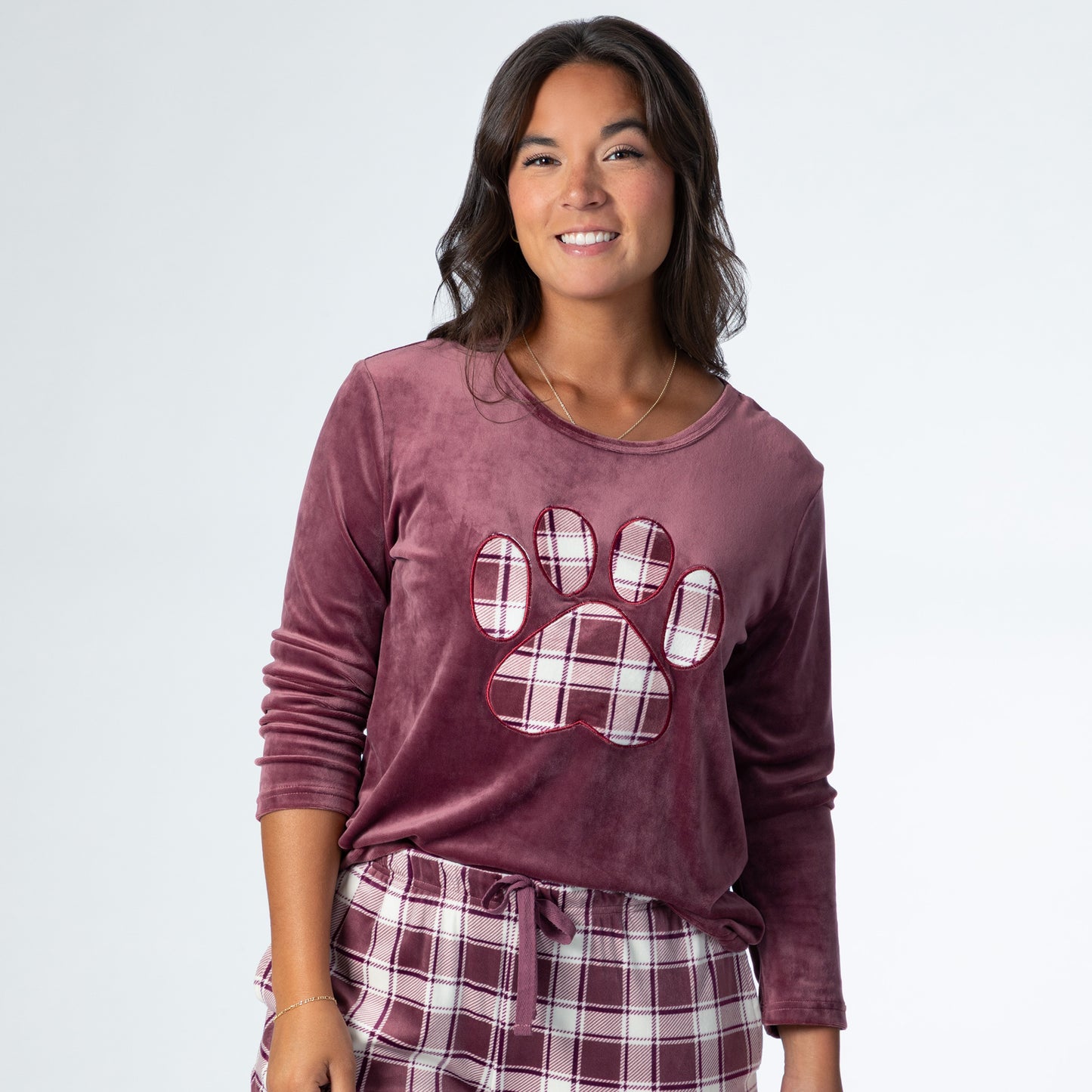 Woman wearing a velour pajama set— the top is a maroon long-sleeve shirt with a large plaid paw print. The pants are the same white and maroon plaid pattern as the paw print on the shirt. The shirt sleeves are rolled up to the elbows and the hem is tucked into the adjustable waistband of the pants.