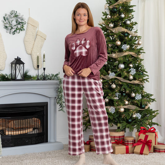 Woman standing in cozy holiday setting. She is wearing a velour pajama set— the top is a maroon long-sleeve shirt with a large plaid paw print. The pants are the same white and maroon plaid pattern as the paw print on the shirt. 