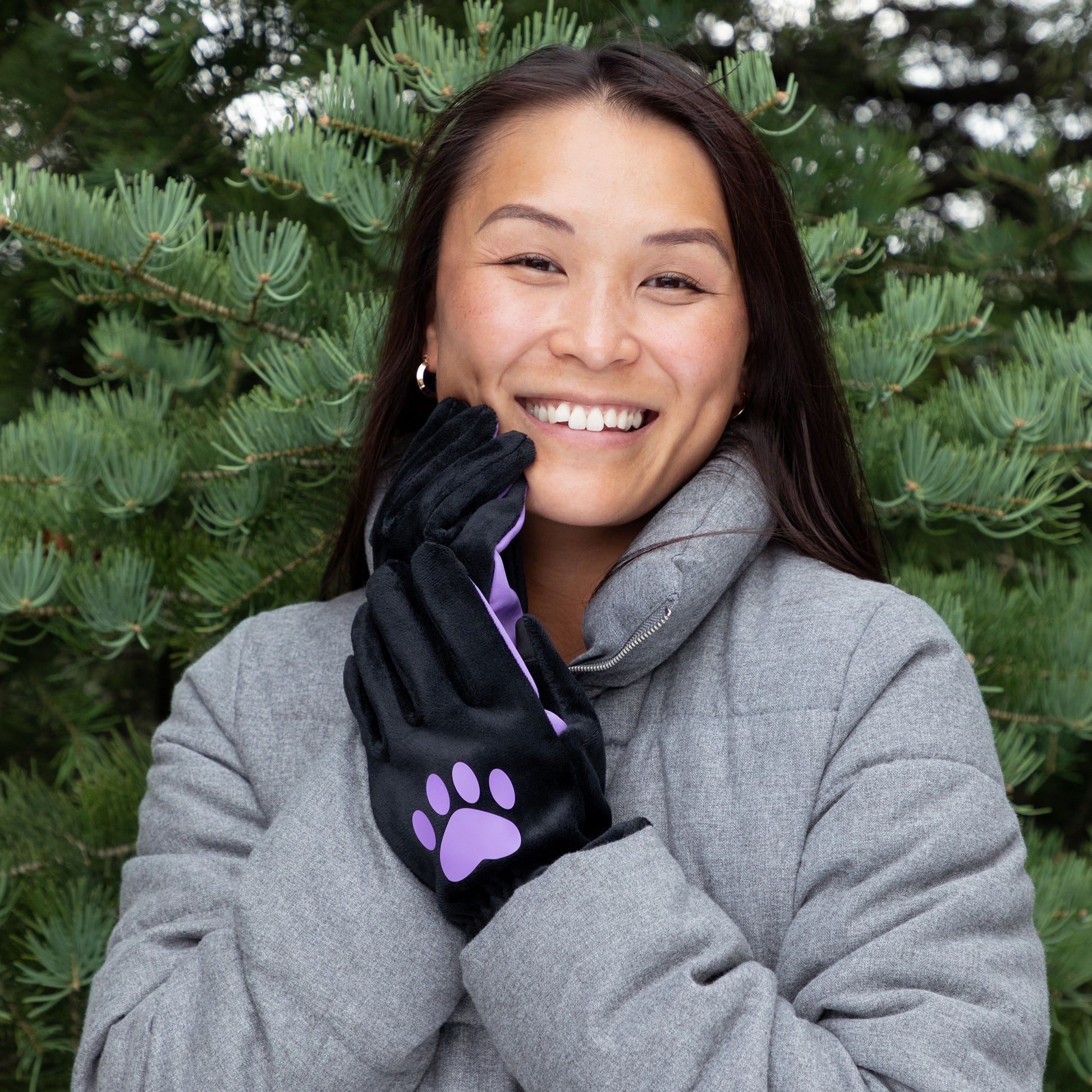 Woman wearing a gray coat and black gloves with a purple paw print, standing in front of green trees.
