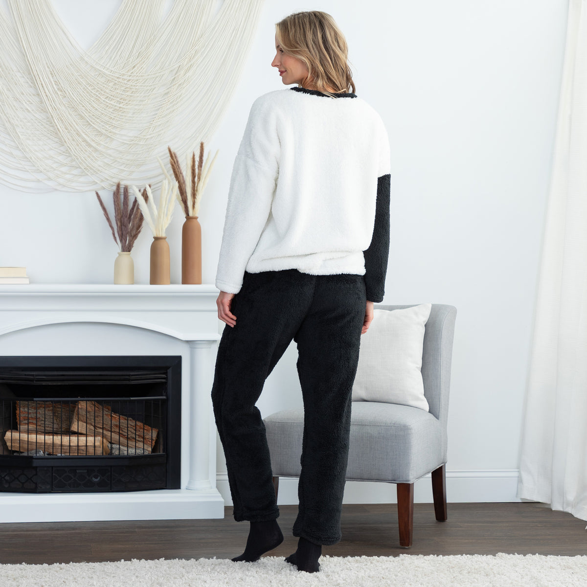 A woman stands with her back to the camera in a cozy living room setting, wearing a white sherpa pullover with black sleeves and black pants.