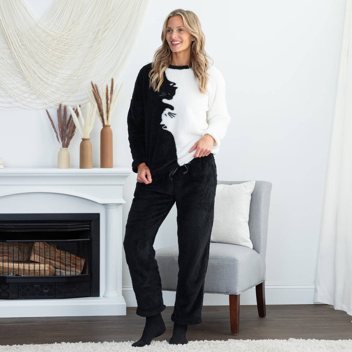 A woman stands in a bright living room wearing a two-piece fuzzy pajama set consisting of a black-and-white asymmetrical sweater and black pants. The background features a white fireplace mantel with decorative vases, a gray armchair, and a white curtain.