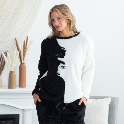A woman wearing a fuzzy black and white long-sleeve top with a cat silhouette design, standing in a bright, modern room with a white mantel and decorative vases.