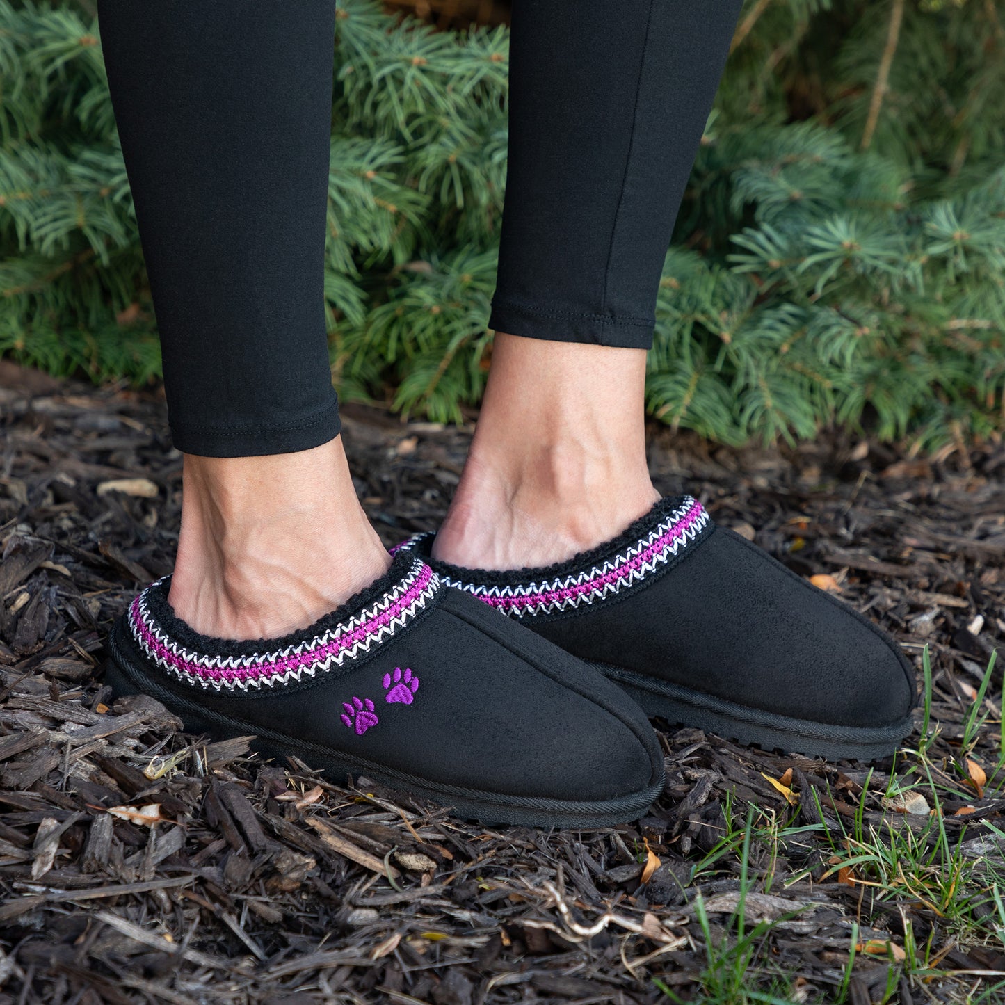 Black slipper shoes with purple accents worn outdoors on a mulched surface.