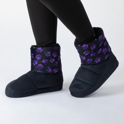 A person wearing black slip-on slipper boots with purple paw print patterns on the cuffs, standing on a white background.