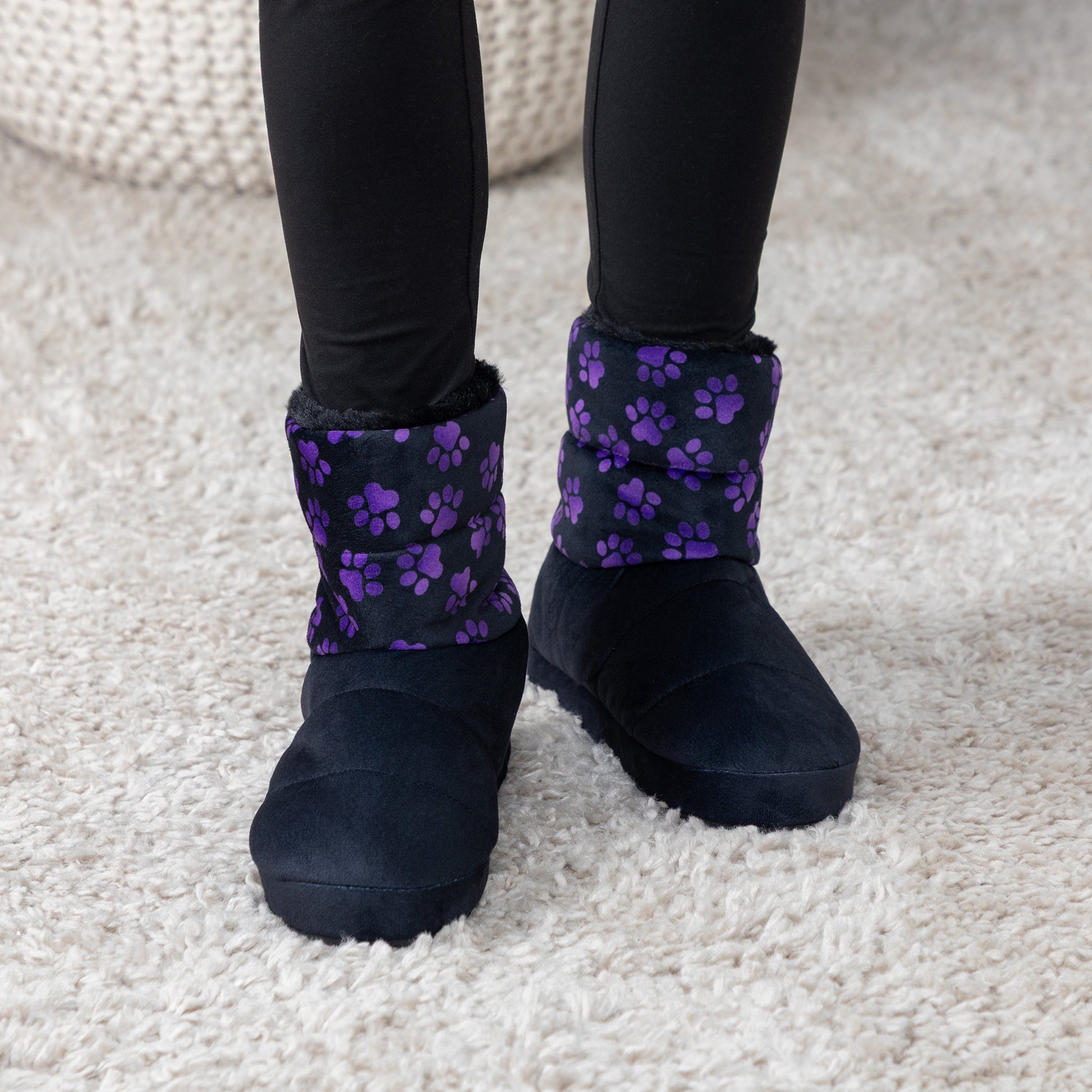 A person wearing black slip-on slipper boots with purple paw print patterns on the cuffs, standing on a beige shag rug.