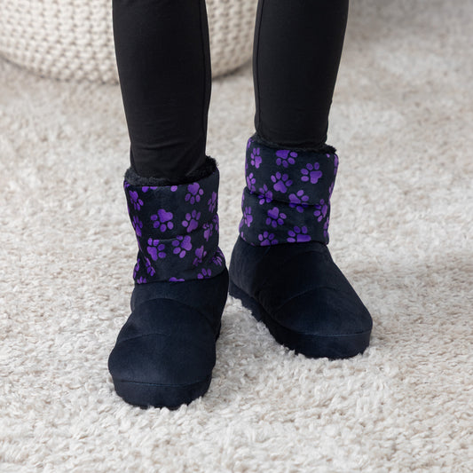 A person wearing black slip-on slipper boots with purple paw print patterns on the cuffs, standing on a beige shag rug.