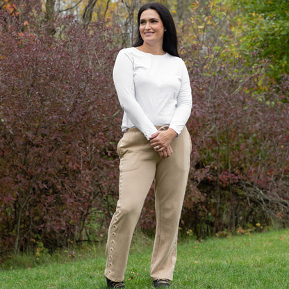 Woman wearing a white long-sleeve top and beige pants standing in a grassy area with trees in the background.