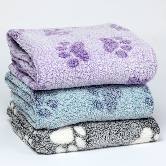 Three folded plush fleece blankets stacked on a white background, each with a purple paw print pattern. The top blanket is lavender, the middle is light blue, and the bottom is gray and white.