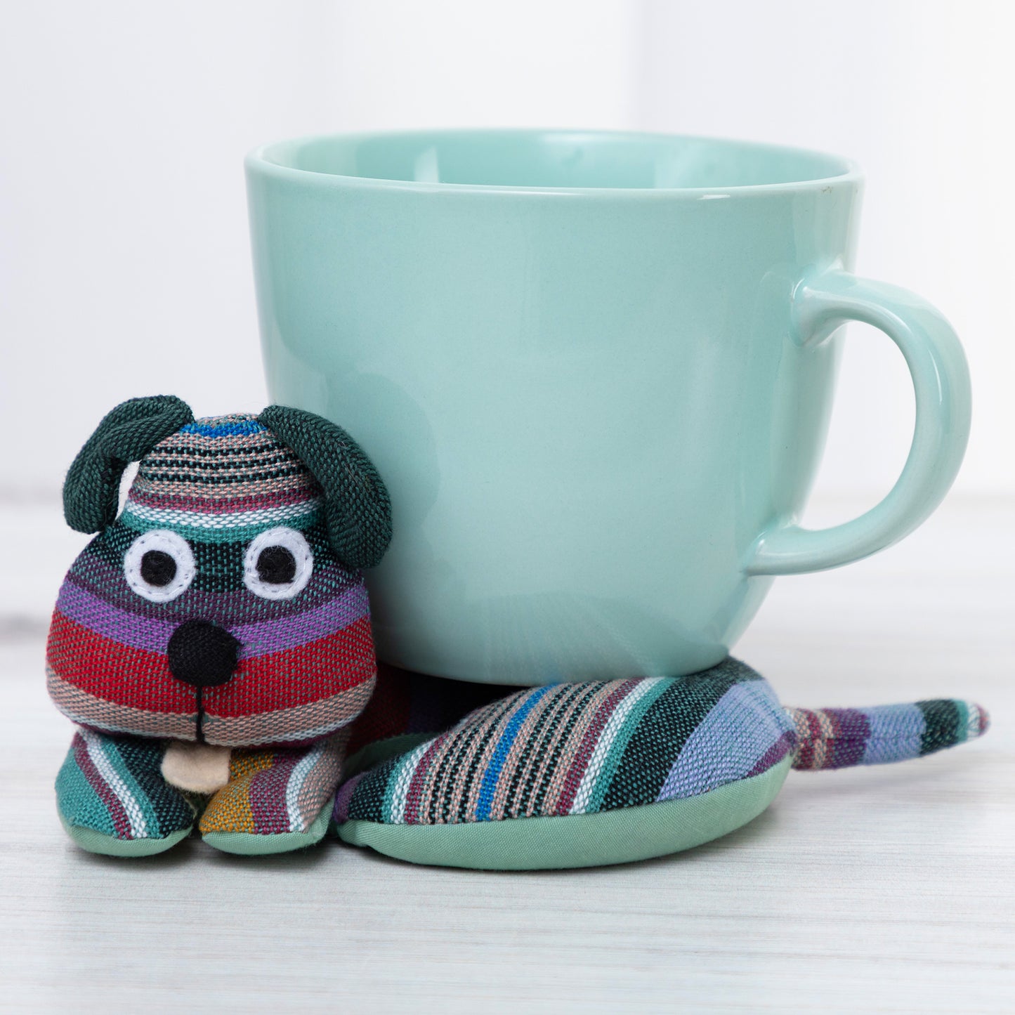 Colorful striped dog-shaped tea cozy next to a light blue mug on a white background
