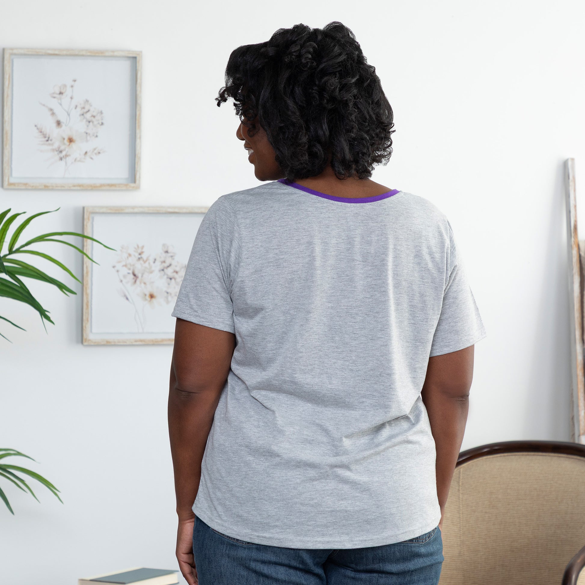 Woman wearing a gray t-shirt with a purple trim in a room with plants and framed pictures on the wall.