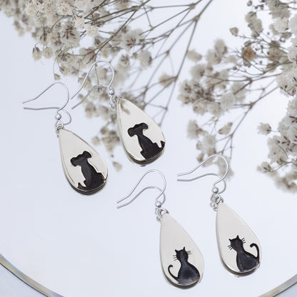 Teardrop-shaped silver earrings with black cat or black dog designs on a white background with dried flowers.