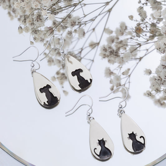 Teardrop-shaped silver earrings with black cat or black dog designs on a white background with dried flowers.