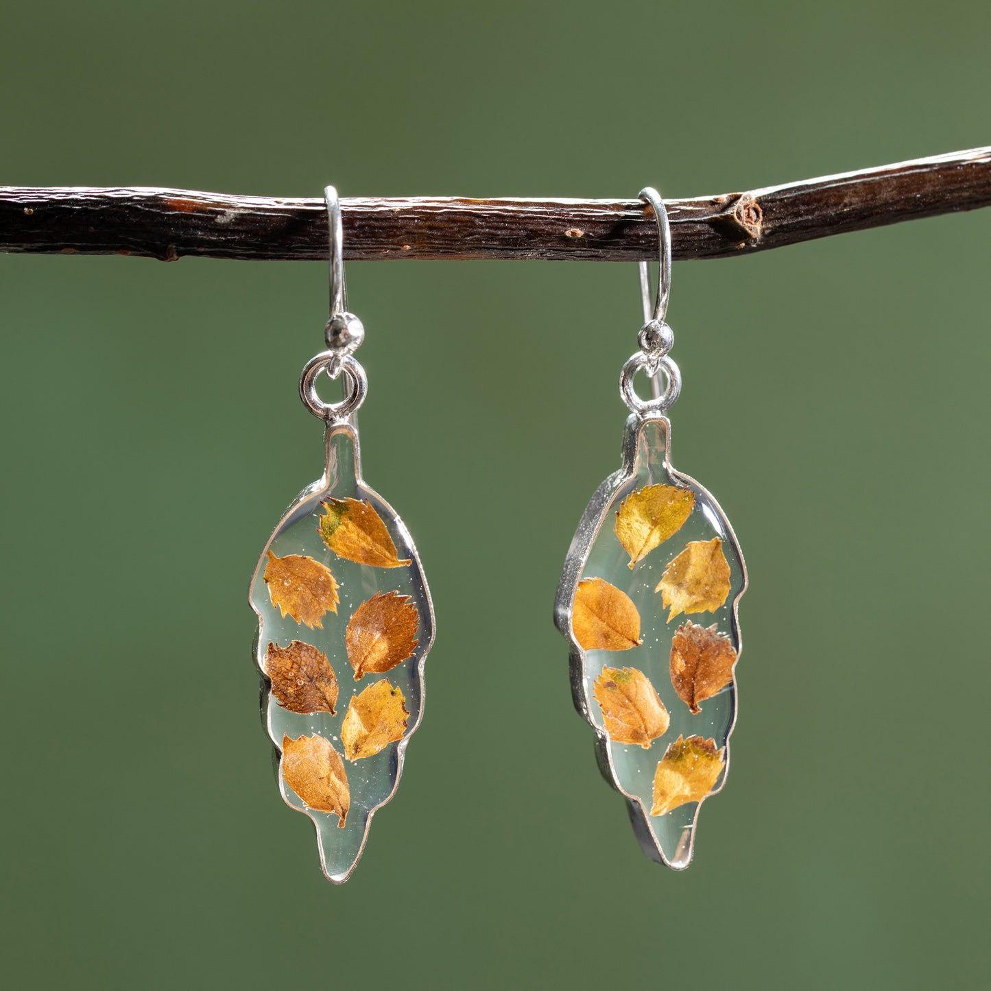 Silver earrings with leaf-shaped designs featuring embedded yellow and brown leaves on a green background.