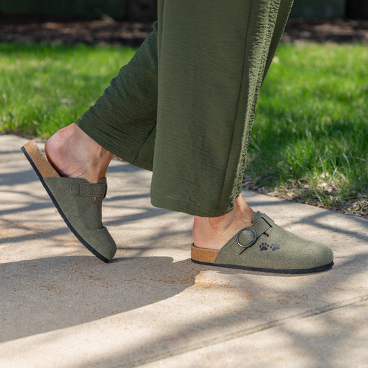Person wearing green slip-on shoes while walking on a sidewalk beside grass.