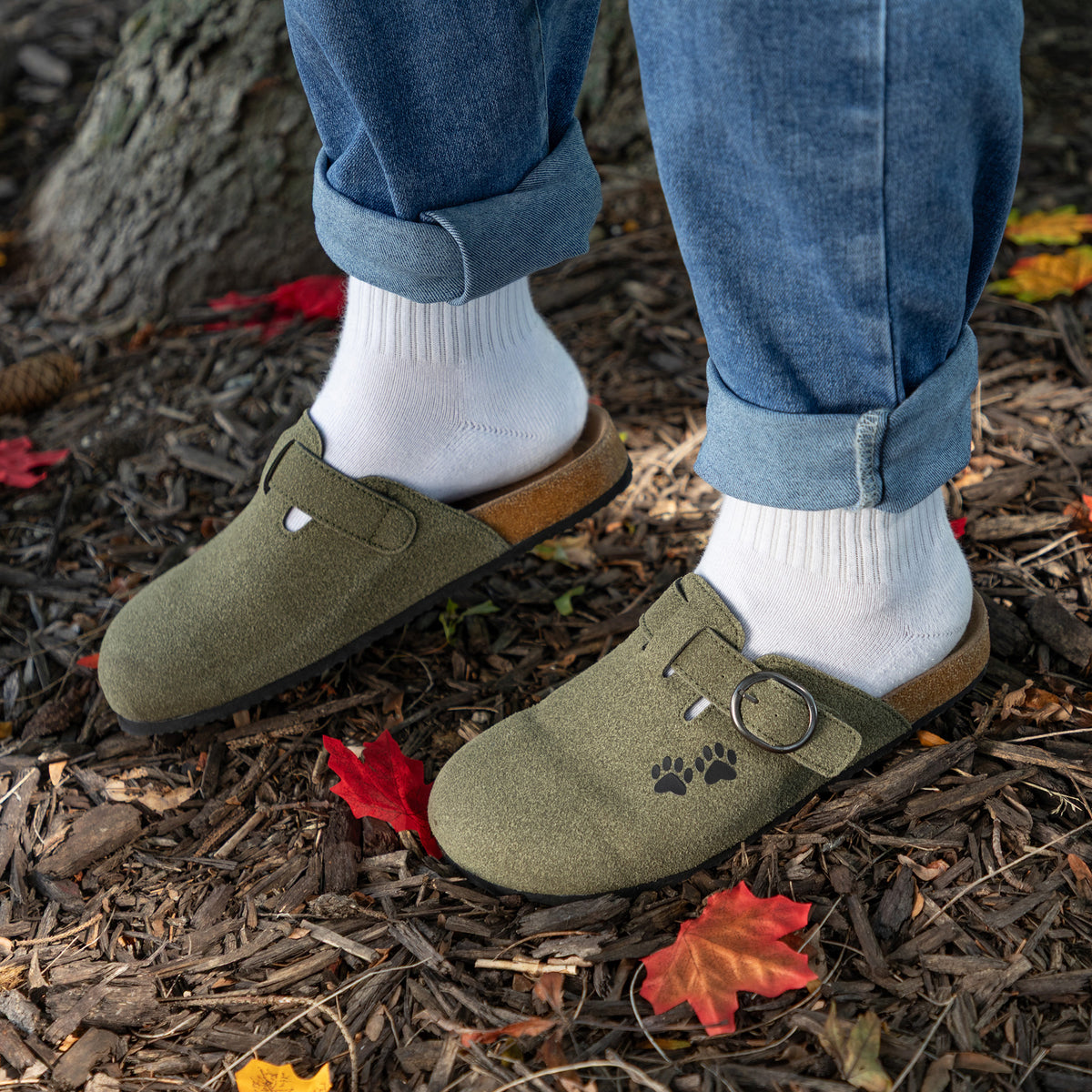 A person wearing olive green clogs with white socks and blue jeans, standing outdoors on a bed of brown wood mulch and fallen red leaves. The clogs have a suede-like finish, a cork midsole, and a small black paw print detail on the side.
