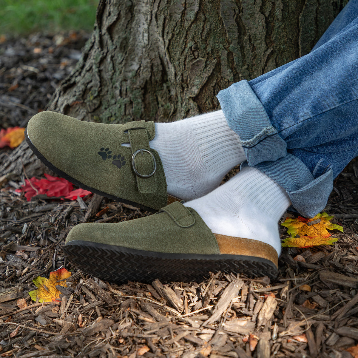 Olive green suede clog slip-on shoes with a buckle strap and paw print logo, worn with white socks and cuffed blue jeans on a forest floor with mulch and fallen leaves.