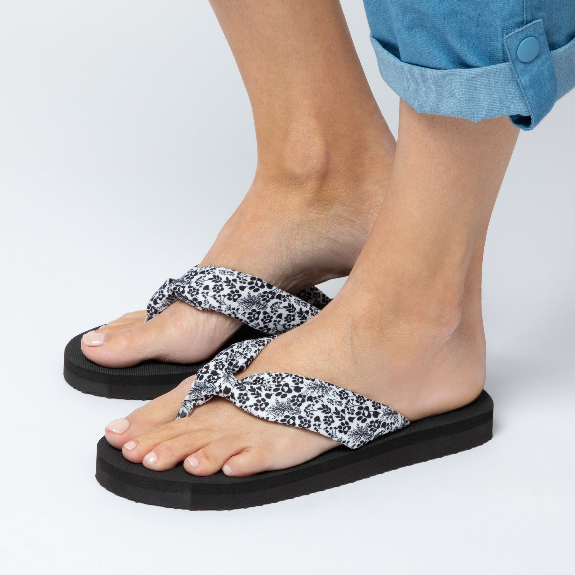 Black flip-flops with white floral straps worn by a person on a light gray background