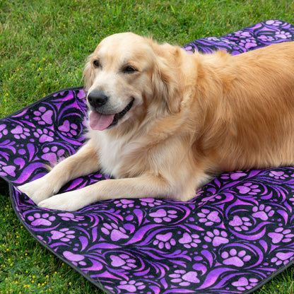 A golden retriever dog lying on a purple and black patterned blanket outdoors on green grass.