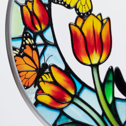 Close-up of a circular stained glass design featuring orange and yellow tulips with butterflies, outlined in silver-toned metal on a white background.