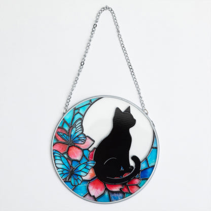 A circular stained-glass-style hanging ornament featuring a black cat silhouette seated in front of a white crescent moon and colorful blue, pink, and teal butterfly and flower motifs, suspended by a silver chain on a white background.