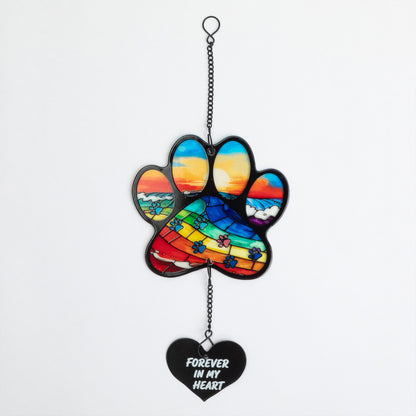 A hanging paw-shaped memorial ornament with a colorful sunset landscape design and a black heart charm below that reads 'FOREVER IN MY HEART' in white text, shown against a plain light background.