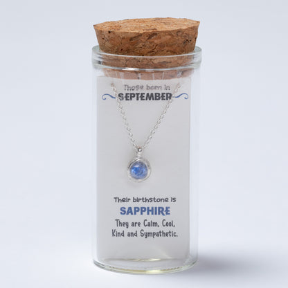 Glass jar with cork lid containing a necklace with a blue gemstone, on a light gray background.