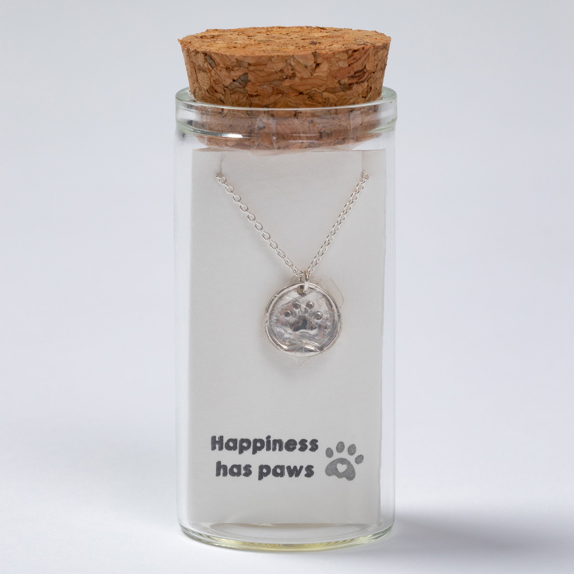 Glass jar with cork lid containing a necklace with a paw print pendant, on a light gray background.