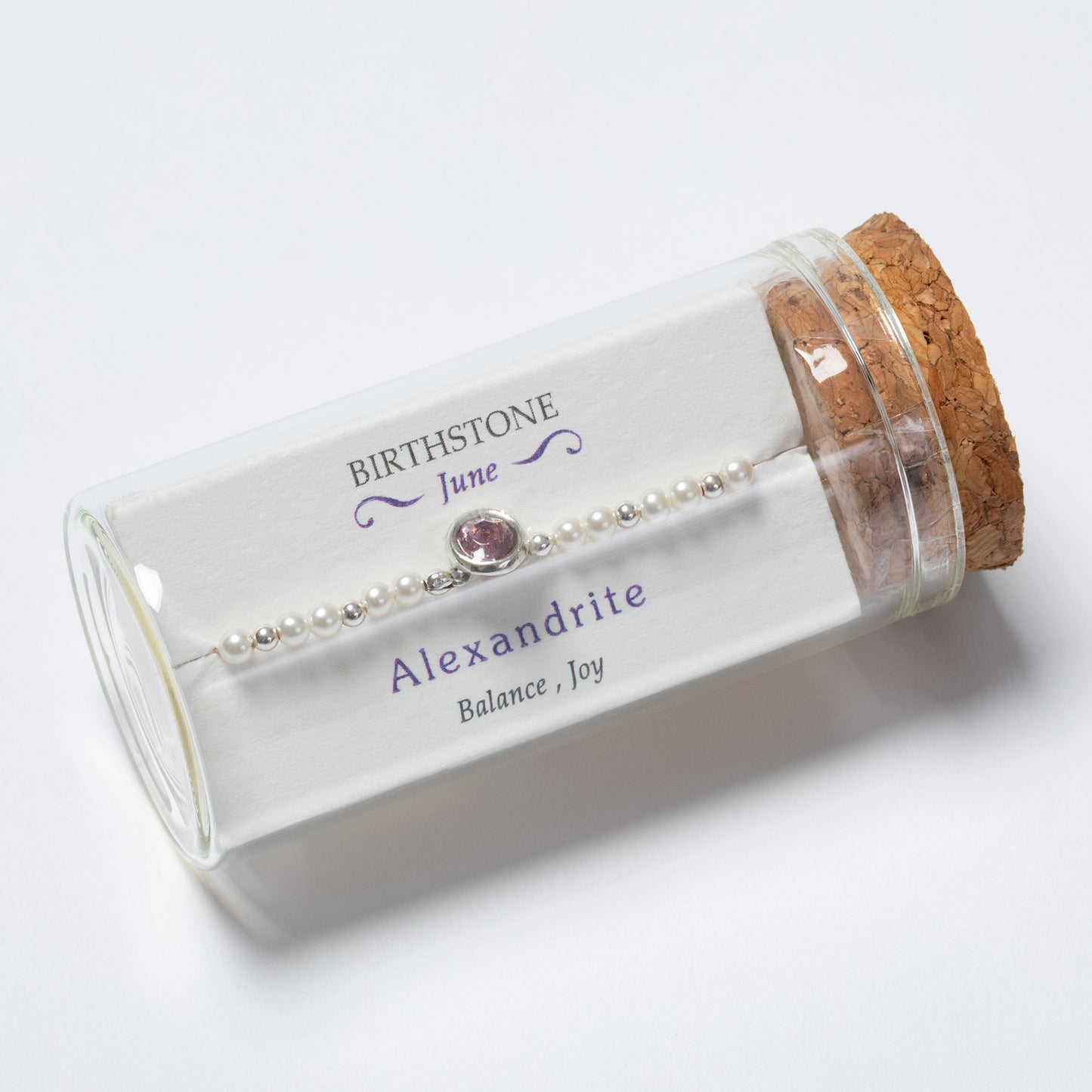 Glass jar with cork lid containing a pearl bracelet with a red gemstone, labeled 'Birthstone June Alexandrite'.