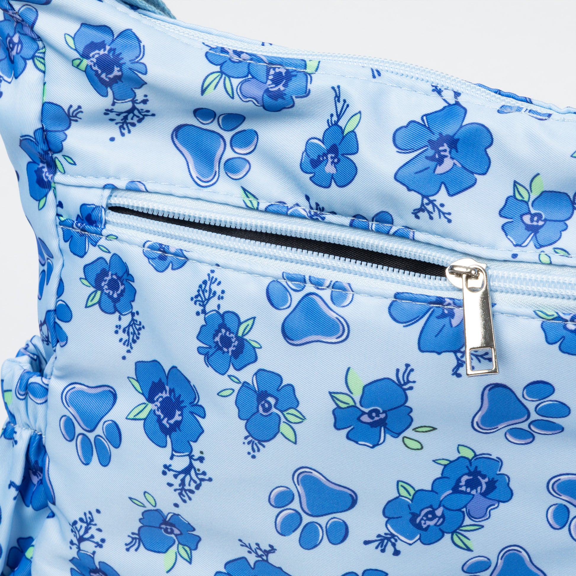 A close-up of a light blue bag or pouch with a blue floral and paw print pattern, featuring a horizontal zipper with a silver pull.