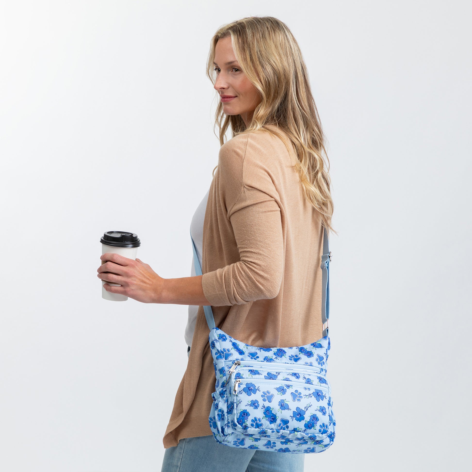 A woman in a tan cardigan and jeans carries a blue and white floral and paw print patterned crossbody bag while holding a coffee cup against a plain light gray background.