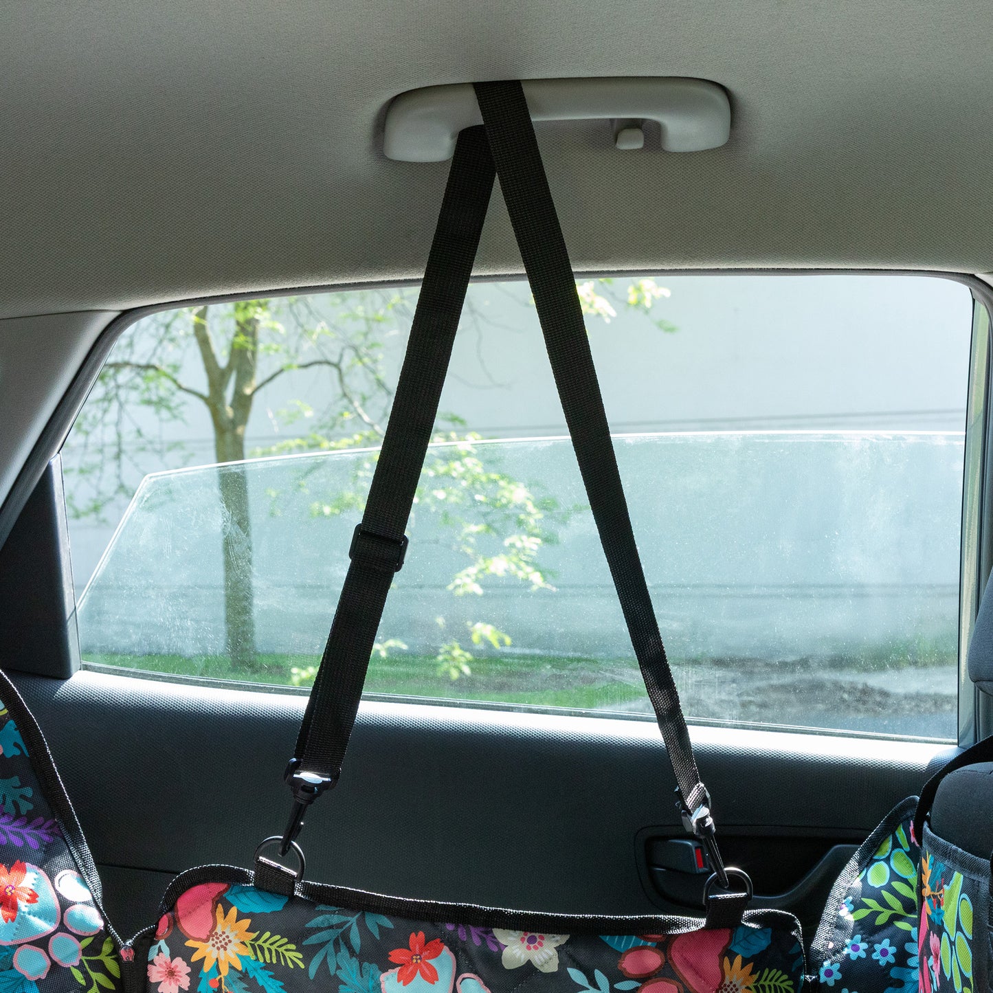 Floral-patterned car seat cover with a black strap over a car seat, with a blurred outdoor background.
