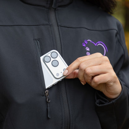 A close-up of a person wearing a black zip-up jacket with a purple heart-shaped paw print logo on the chest. The person is holding a silver smartphone in their hand, showing how the phone fits into a small zippered pocket on the jacket.