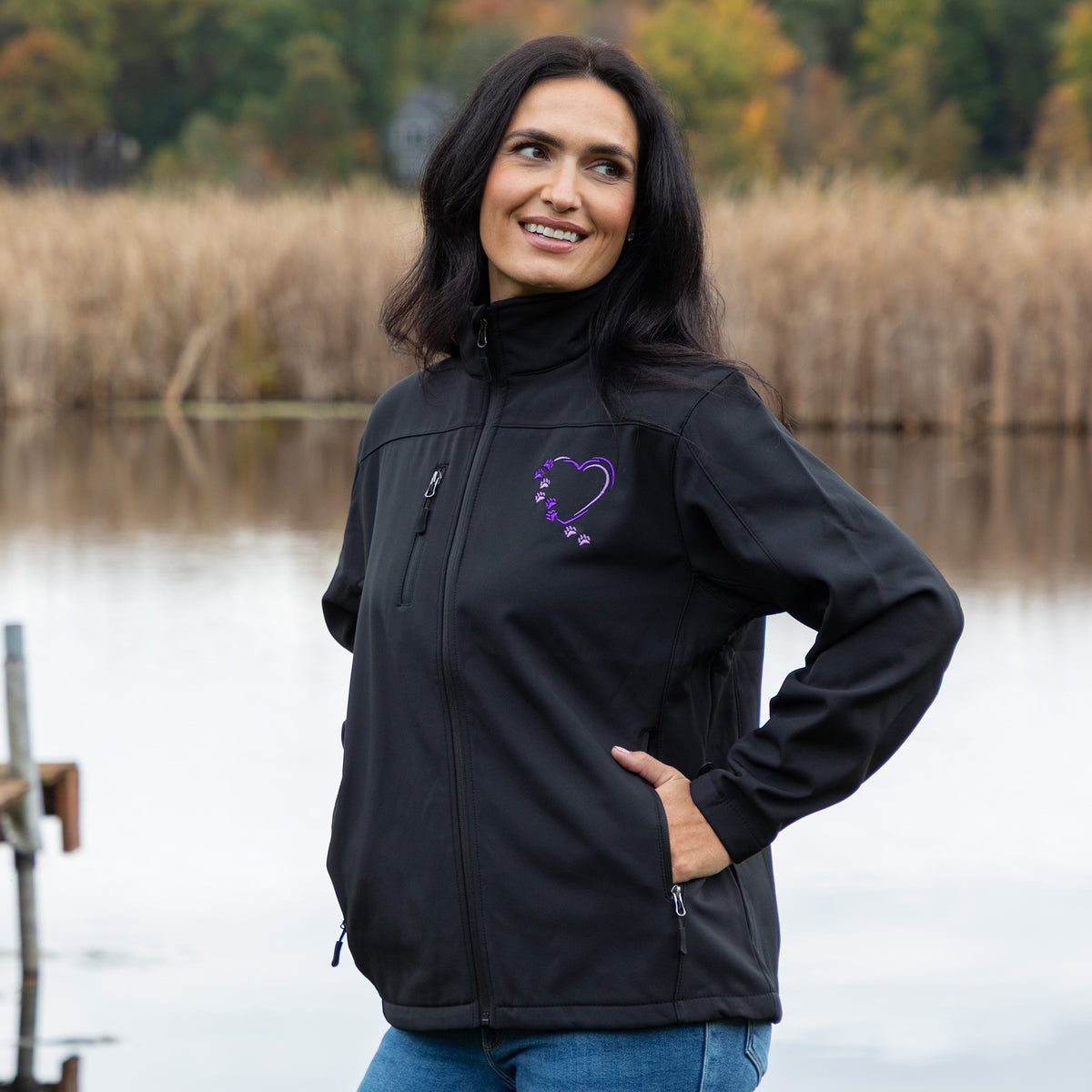A woman wearing a black softshell jacket with a purple embroidered heart logo on the left chest, standing outdoors near a body of water with reeds in the background.