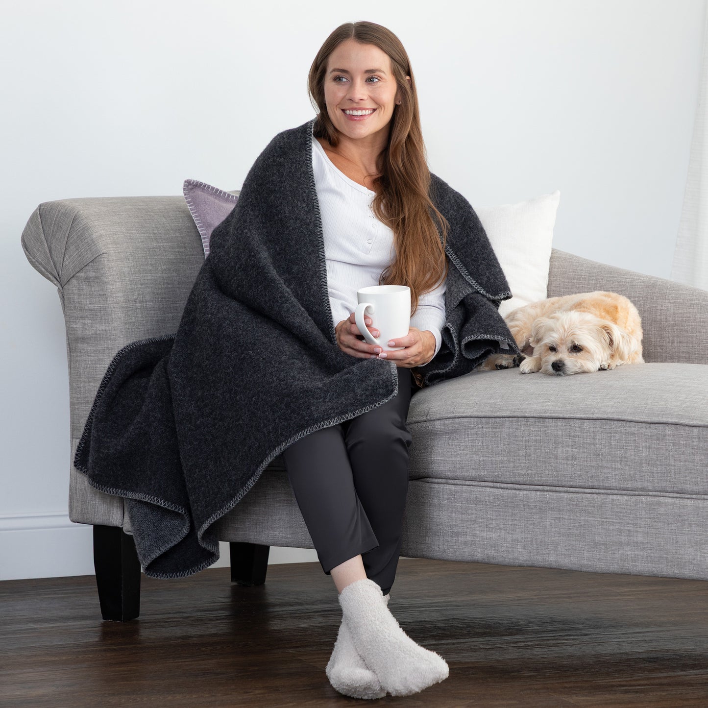 A woman sits on a gray sofa wrapped in a dark charcoal heather throw blanket, holding a white mug, with a small light-colored dog resting on the sofa beside her. The setting is a cozy living room with a light gray wall and dark wood floor.