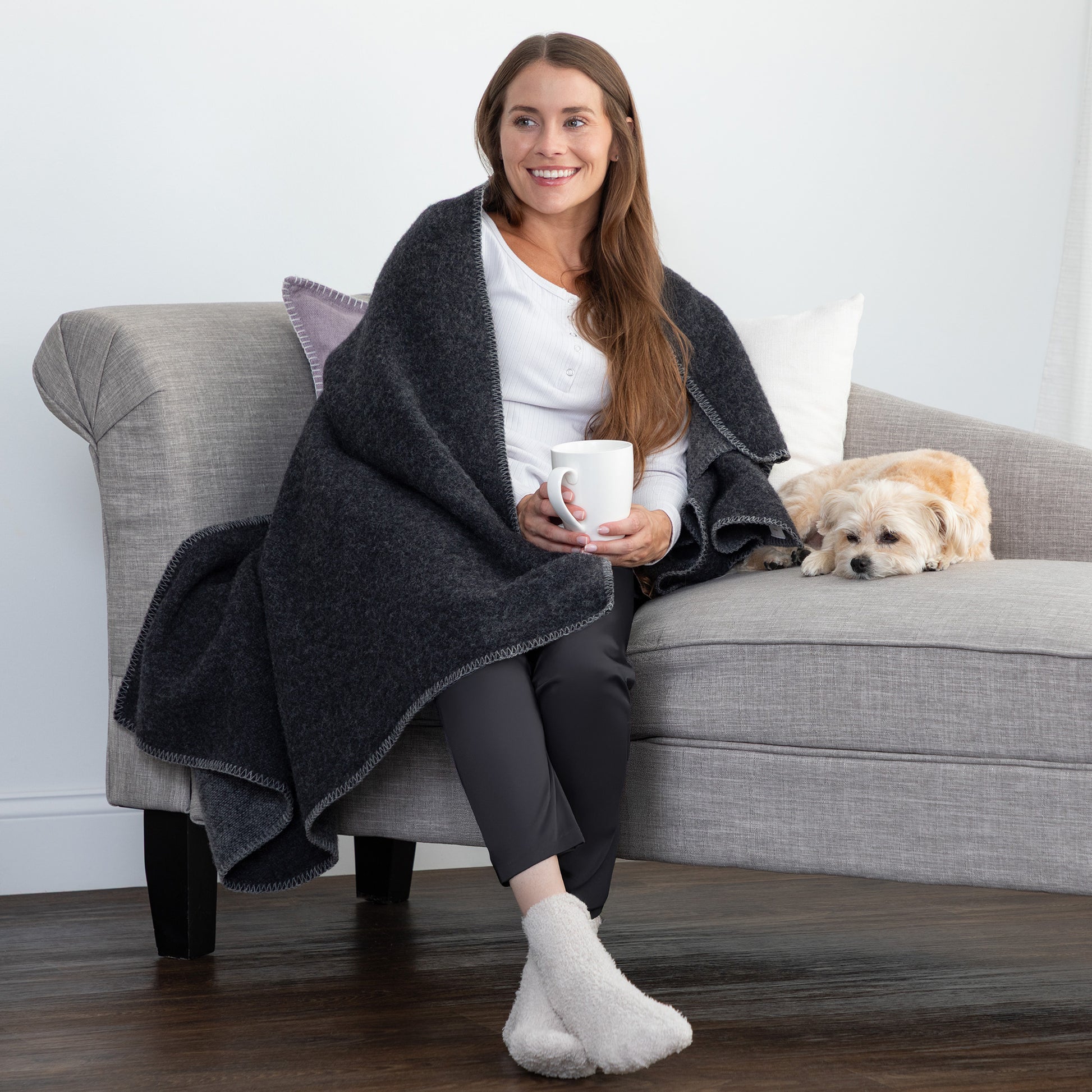 A woman sits on a gray sofa wrapped in a dark charcoal heather throw blanket, holding a white mug, with a small light-colored dog resting on the sofa beside her. The setting is a cozy living room with a light gray wall and dark wood floor.