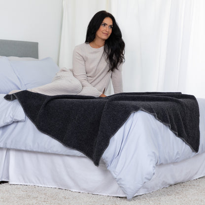 A woman sits on a bed with white bedding, draped over her lap a dark charcoal gray knit blanket or throw. She wears a light beige ribbed long-sleeve top in a bright bedroom with white curtains and a gray headboard.