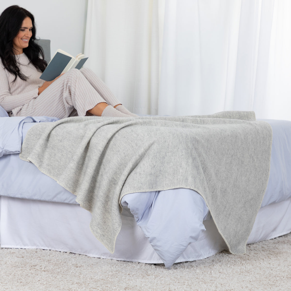 A woman in light pink pajamas sits on a bed reading a book, with a light gray heathered knit blanket draped over the bed in a bright, neutral bedroom setting.