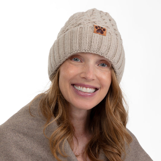 A smiling woman wearing a beige cable-knit beanie with a small brown paw-print patch on the front, against a plain white background.