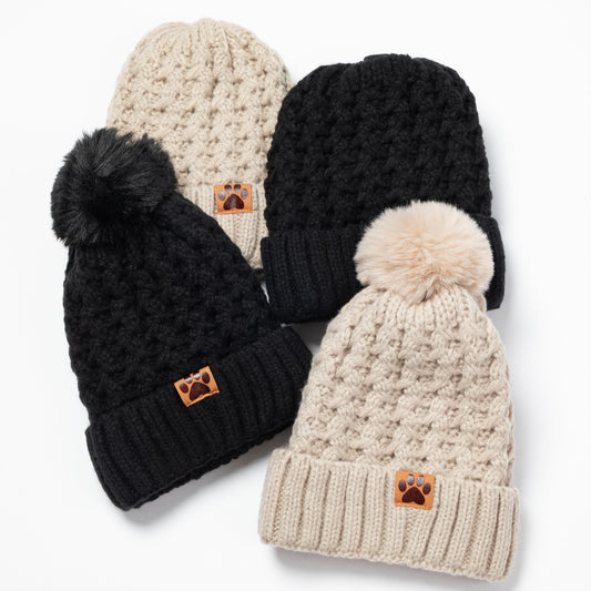 Four knit pom-pom beanies arranged on a white background, including two black and two cream/beige versions, each with a small brown paw-print patch on the cuff.