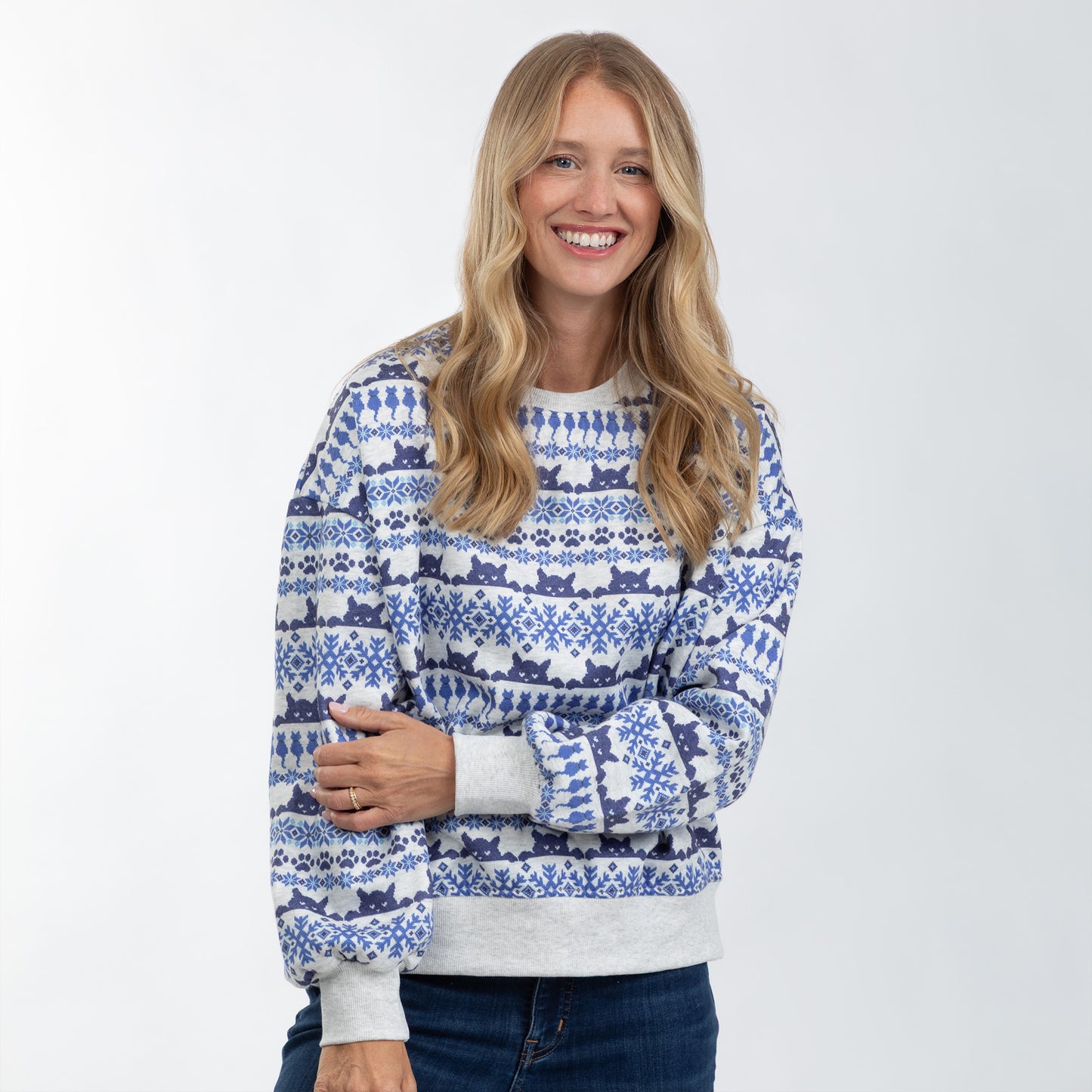A smiling woman wearing a blue and white patterned crewneck sweater with gray ribbed trim, paired with dark blue jeans, against a plain light gray studio background.