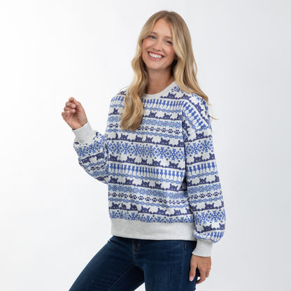 A woman wearing a blue and white patterned crewneck sweater with cat silhouettes and snowflake motifs, paired with dark blue jeans, photographed against a clean white studio background.