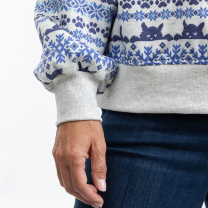 Close-up of a person wearing a light gray crewneck sweater with a blue and navy Nordic pattern, paired with dark blue jeans against a white background.