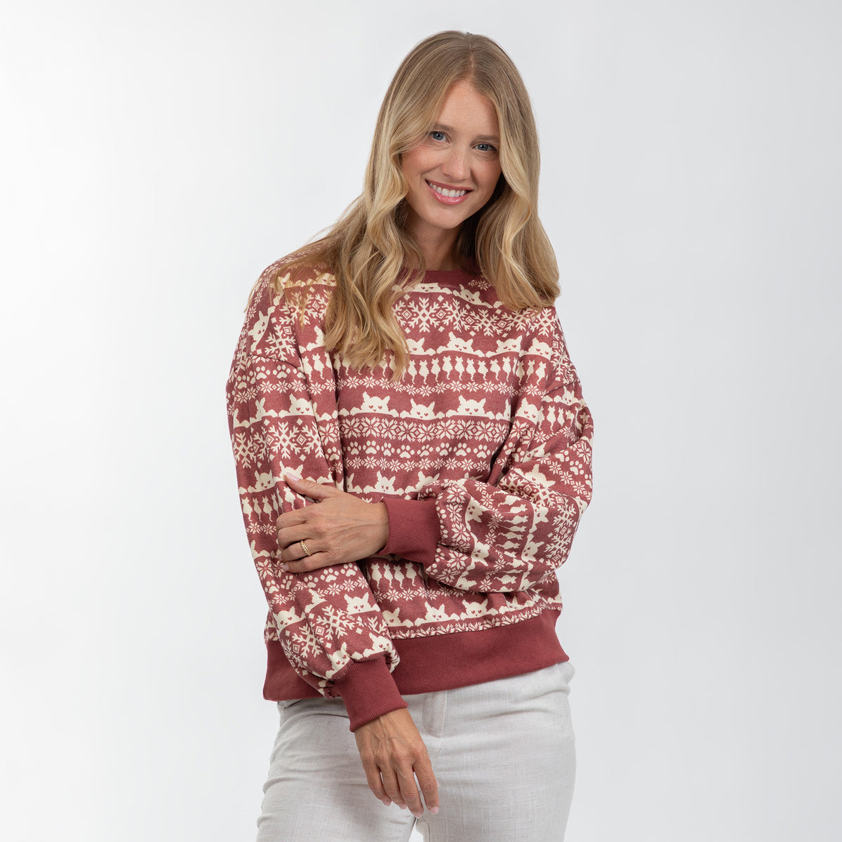 A woman modeling a long-sleeve crewneck sweater with a festive Fair Isle pattern in rust red and cream, paired with light gray pants, against a plain light gray background.