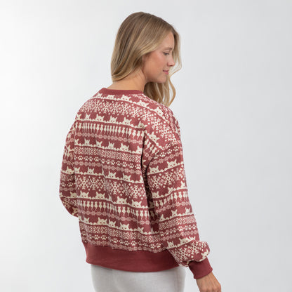 A woman wearing a rust red and cream knit sweater with a repeating cat-themed fair isle pattern, shown from a side-back angle against a light gray studio background.