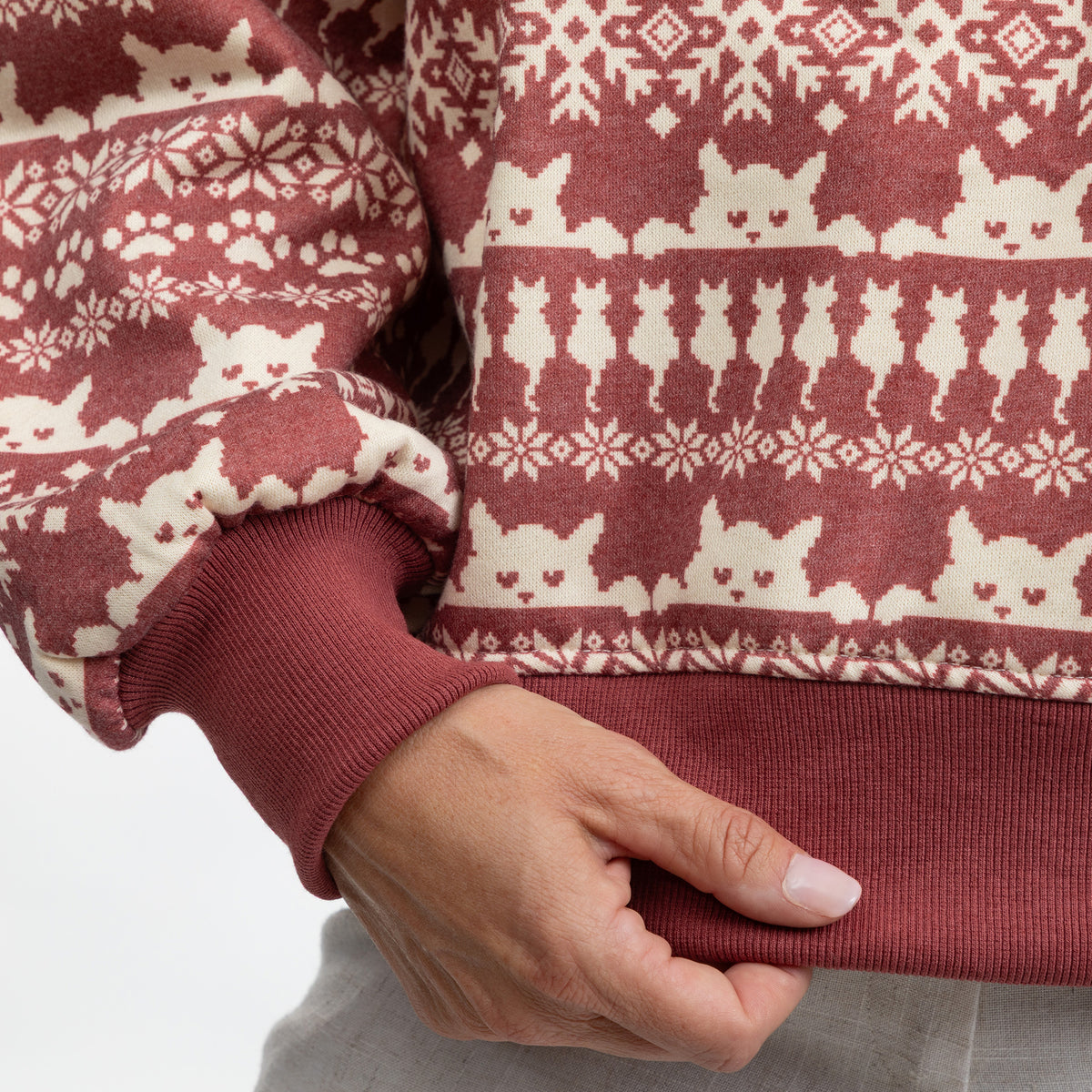 Close-up of a person wearing a ribbed knit sweater with a rust red and cream cat-themed Fair Isle pattern, showing the hand and ribbed cuffs.