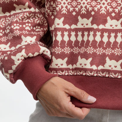 Close-up of a person wearing a ribbed knit sweater with a rust red and cream cat-themed Fair Isle pattern, showing the hand and ribbed cuffs.