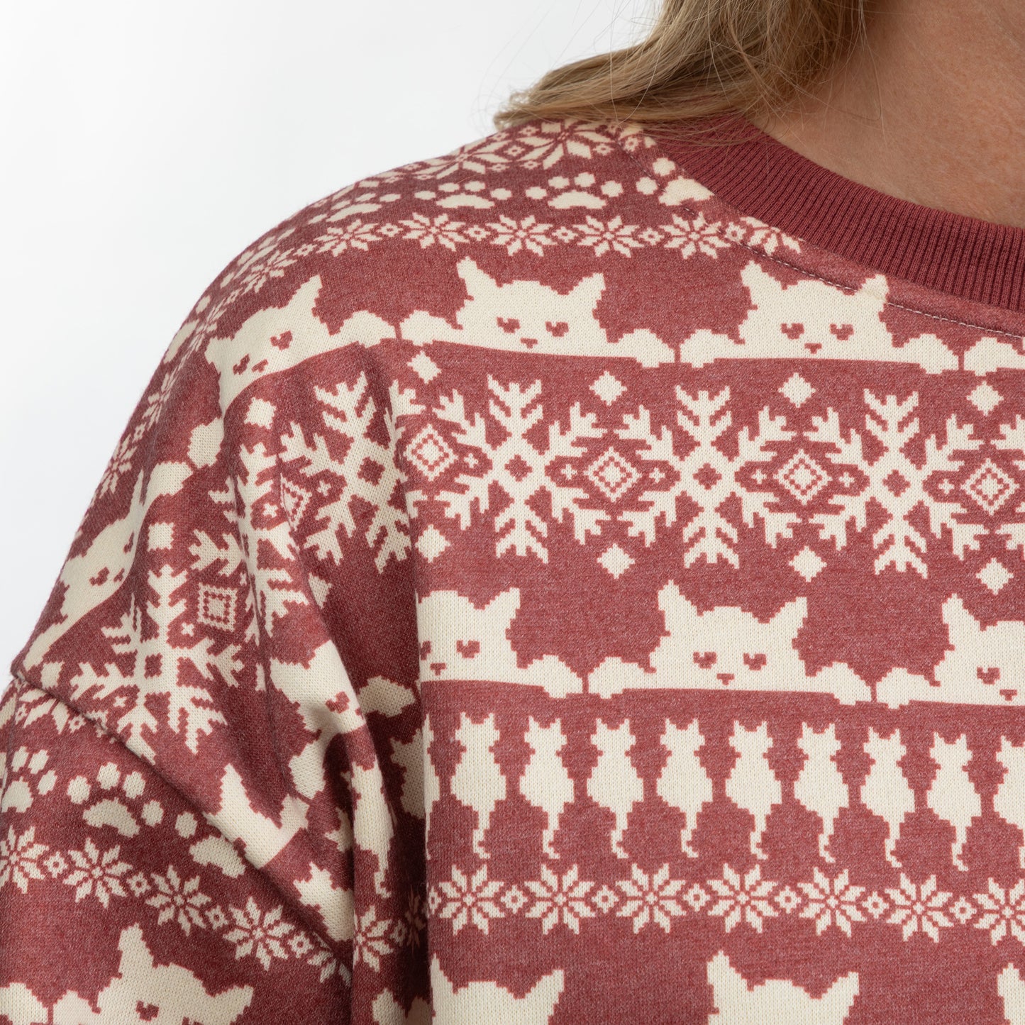 A close-up of a person wearing a festive crewneck sweater with a maroon and cream cat-themed Fair Isle pattern, including snowflakes, hearts, and cat silhouettes.